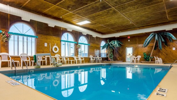 Una piscina interior con agua cristalina, rodeada de sillas y mesas blancas, amplios ventanales y paredes de madera.