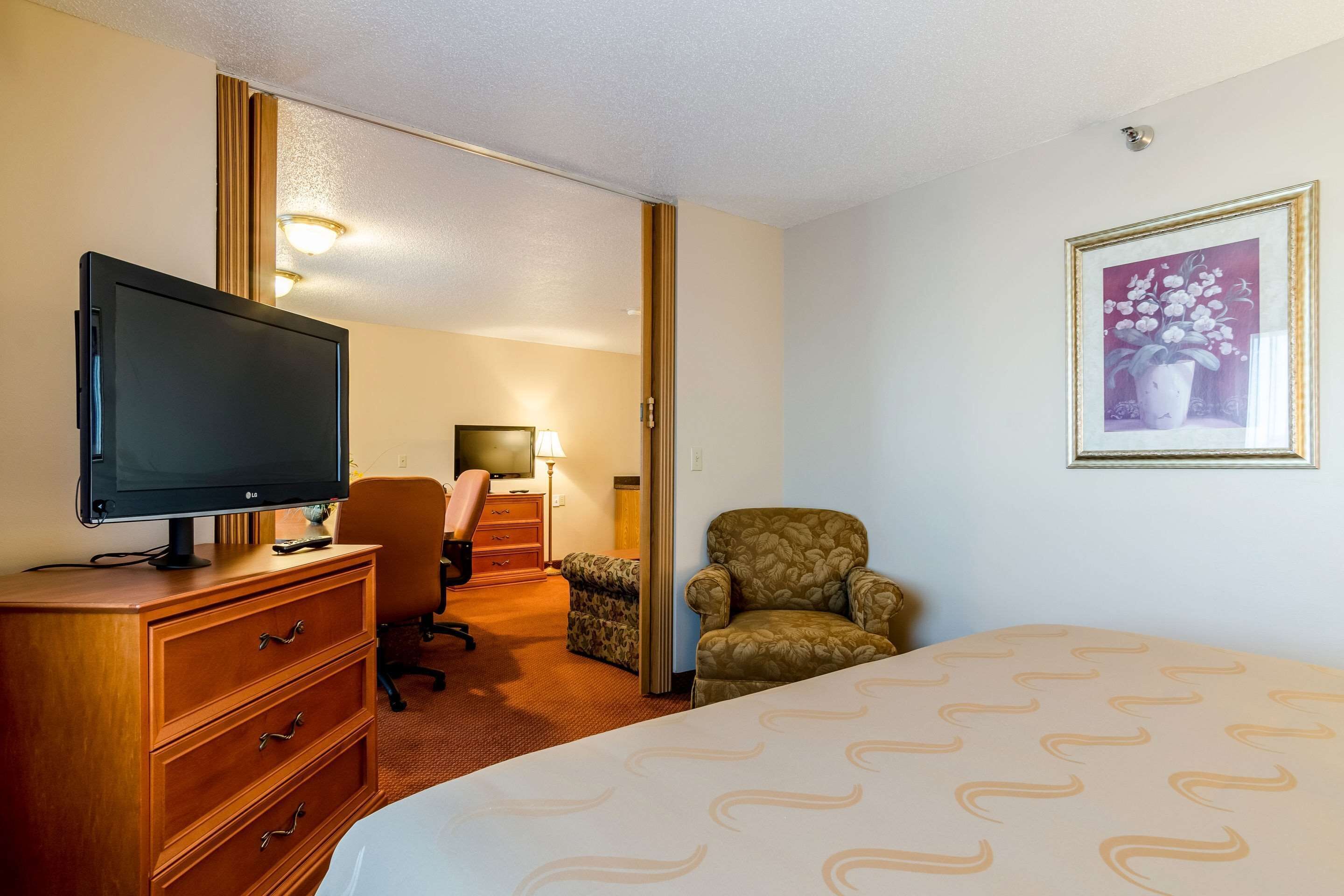 Una habitación de hotel con una cama, un televisor sobre un tocador de madera, una silla, un escritorio con una lámpara y un cuadro en la pared.