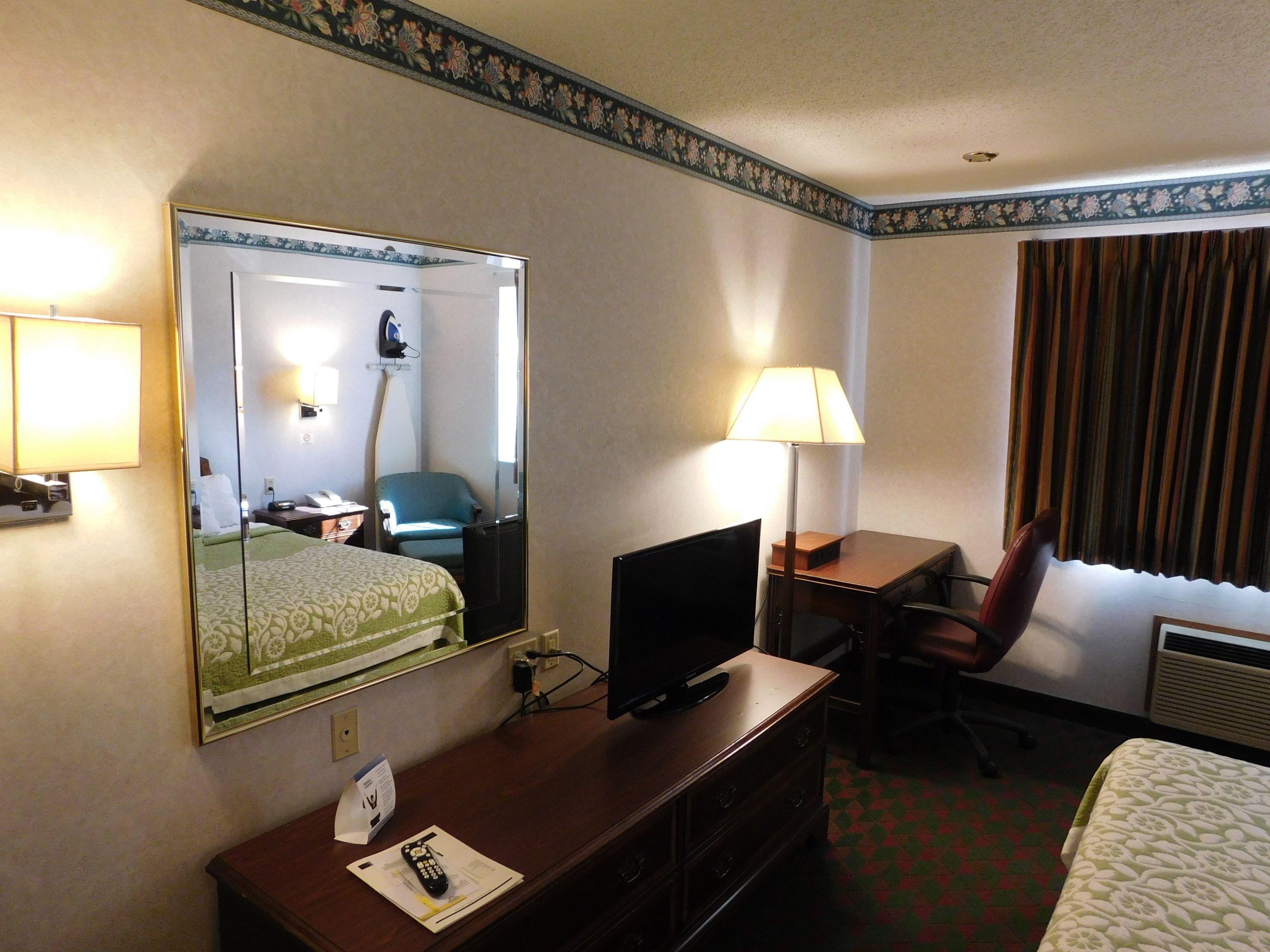A hotel room with a bed, a desk with a computer, a chair, a mirror, and a lamp.