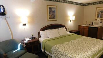 Una habitación de hotel con cama, una silla, una mesita de noche, una lámpara, microondas y refrigerador.