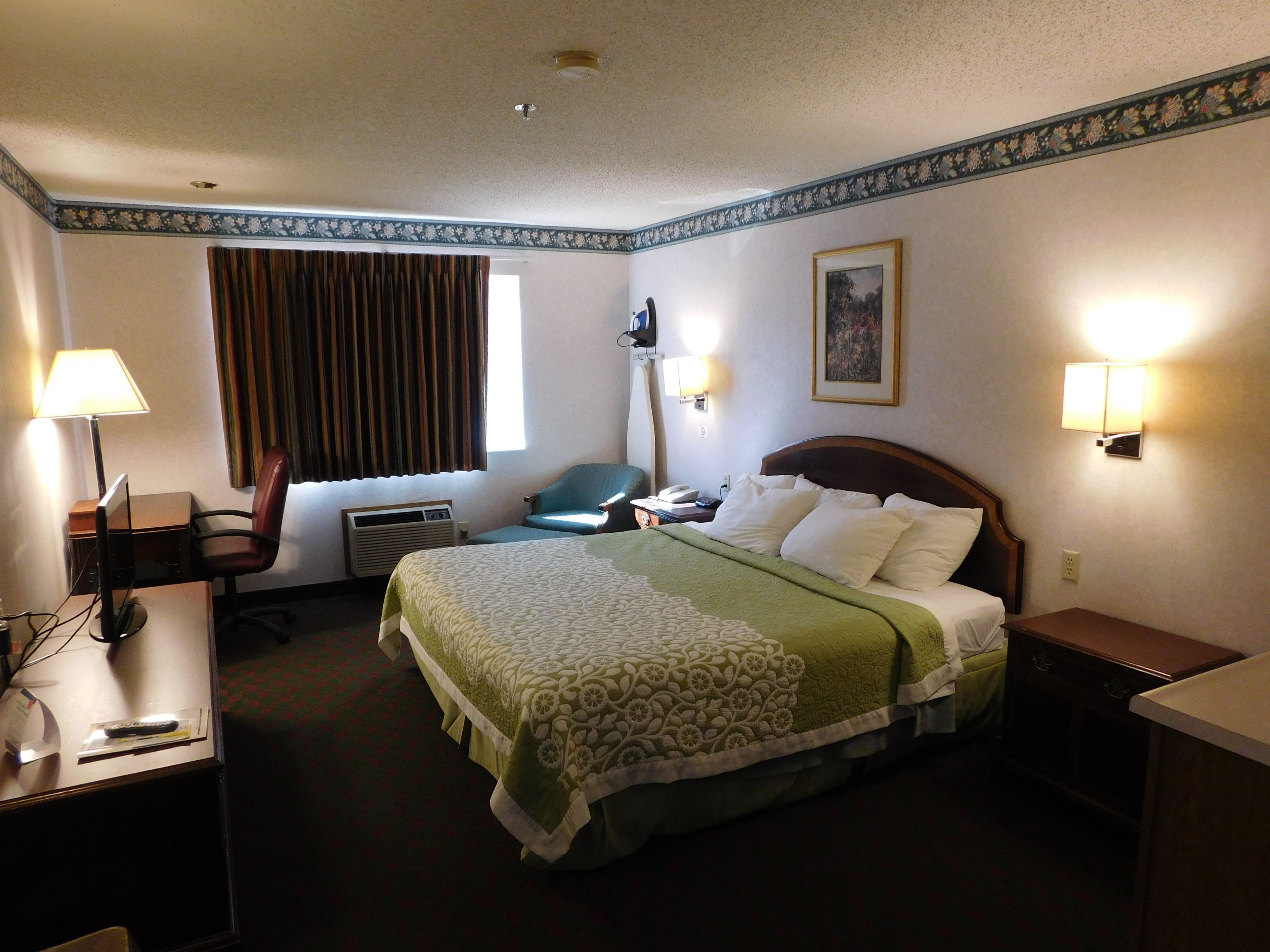 A hotel room with a bed, a desk, a chair, a desk lamp, a wall-mounted lamp, a painting, and a window with curtains.