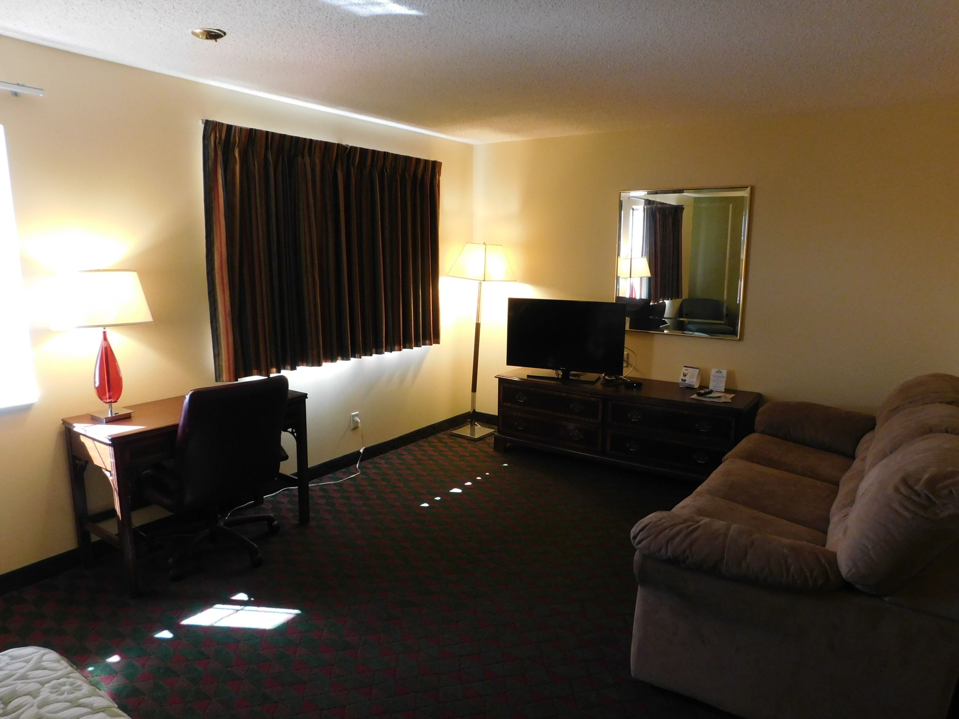A hotel room with a desk, chair, television, sofa, and a mirror.