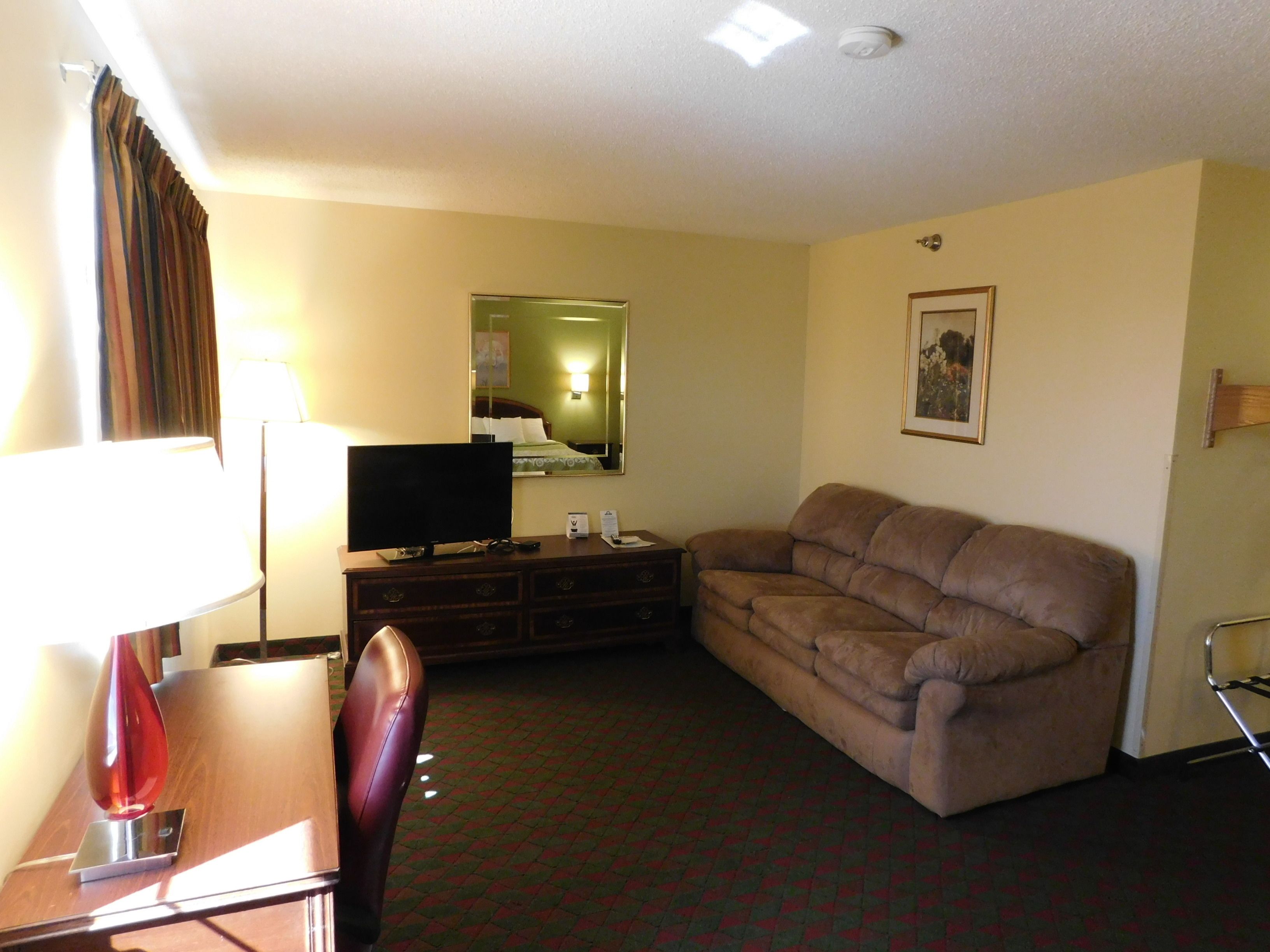 A hotel room with a sofa, a TV on a wooden stand, a desk with a lamp, a chair, and a patterned carpet.