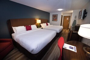 Two single beds with white linens and red accent pillows, a wooden headboard, and a desk with a lamp.
