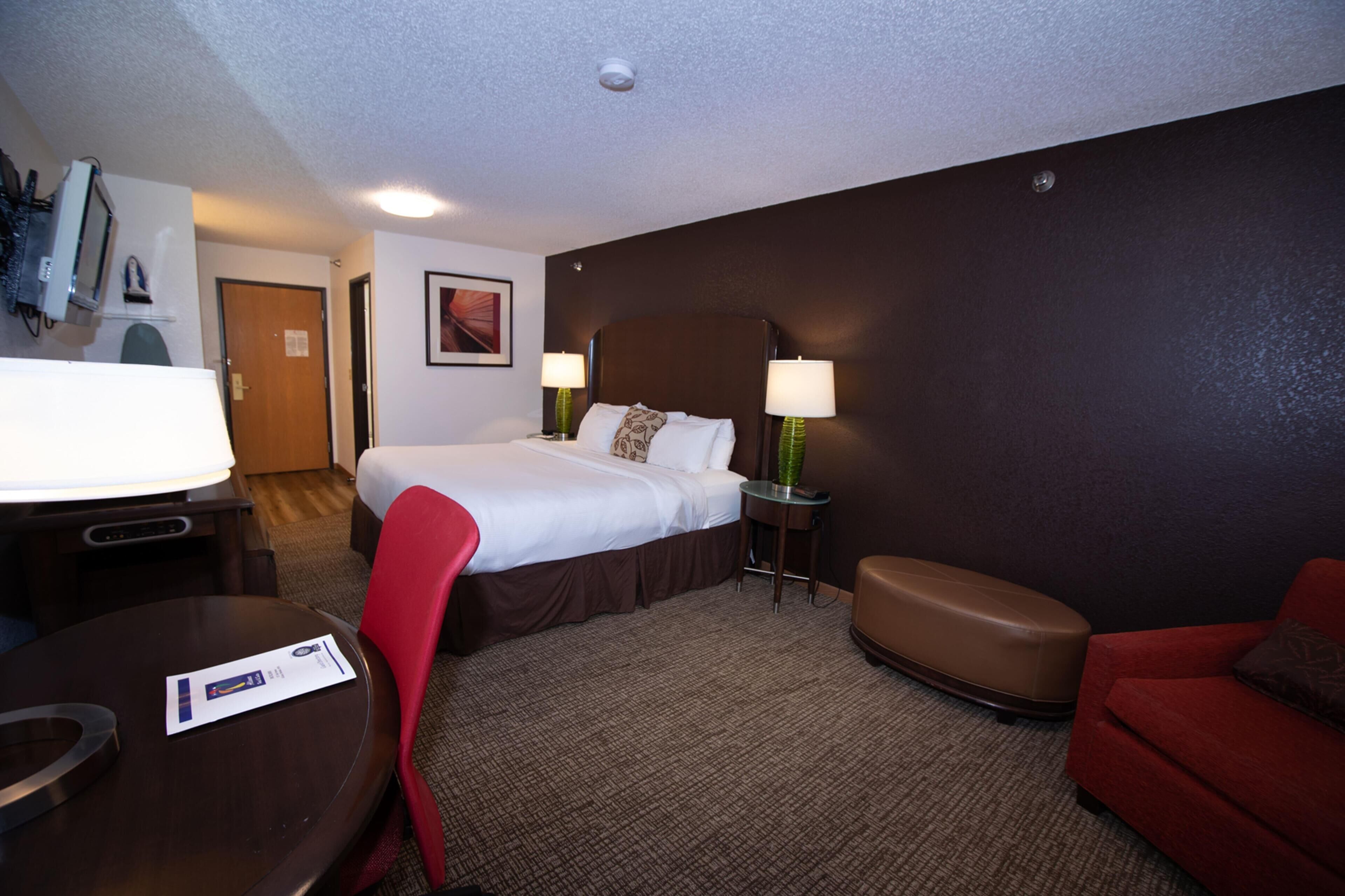 A hotel room with a bed, two bedside tables with lamps, a red chair, a brown ottoman, and a small table with a menu.