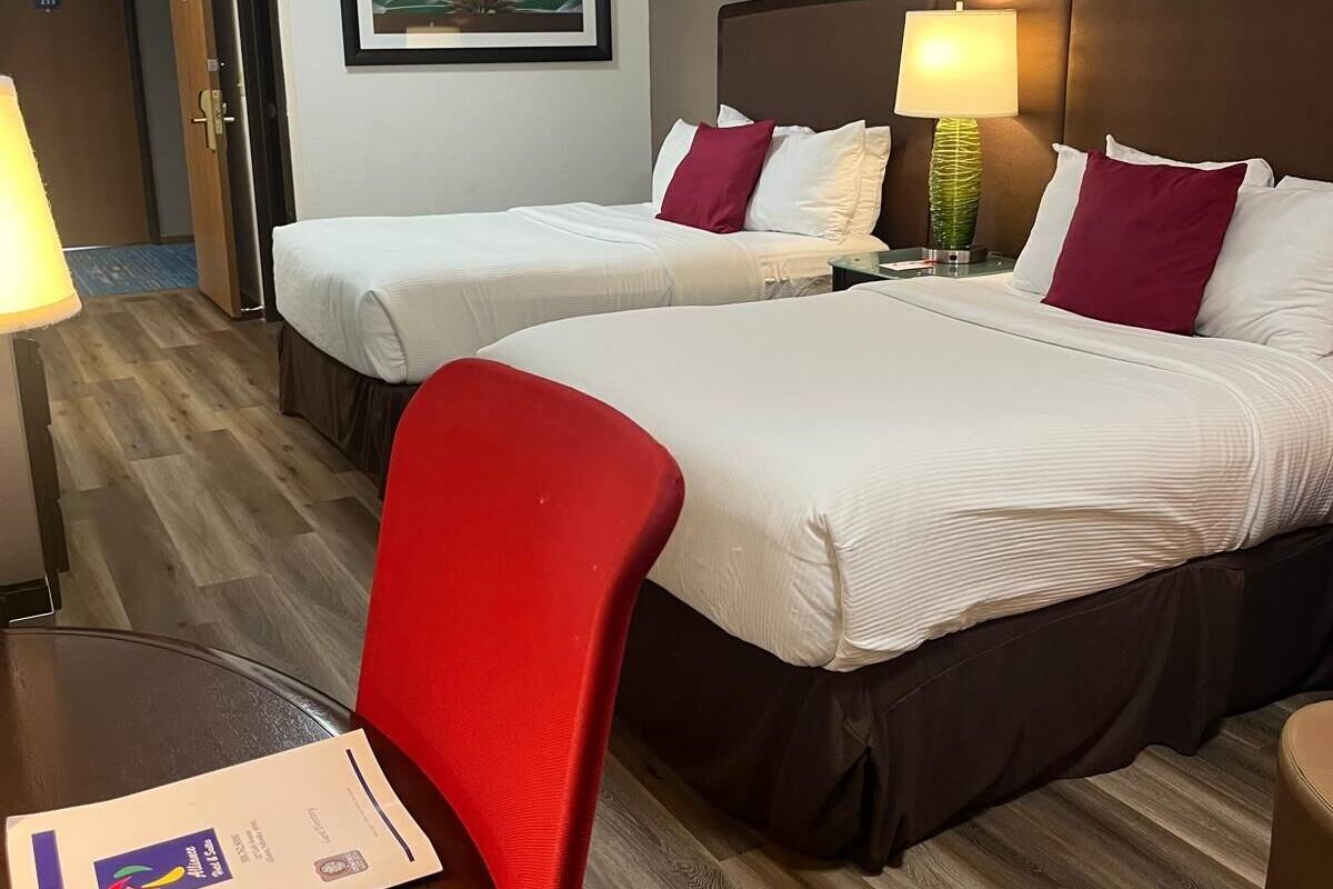 A hotel room with two beds, a red chair, a table with a menu, and a framed plant on the wall.