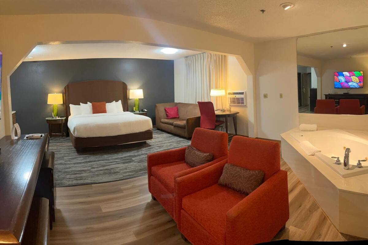 A hotel room with a large bed, a sitting area with two armchairs, a desk, a TV, and a bathtub.