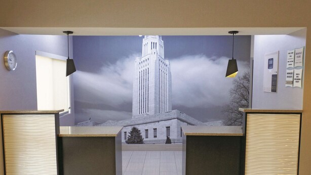 A reception area with a large mural of a building, two reception desks, a clock, and a bulletin board. - La Quinta Inn by Wyndham Lincoln, Nebraska (Lincoln)