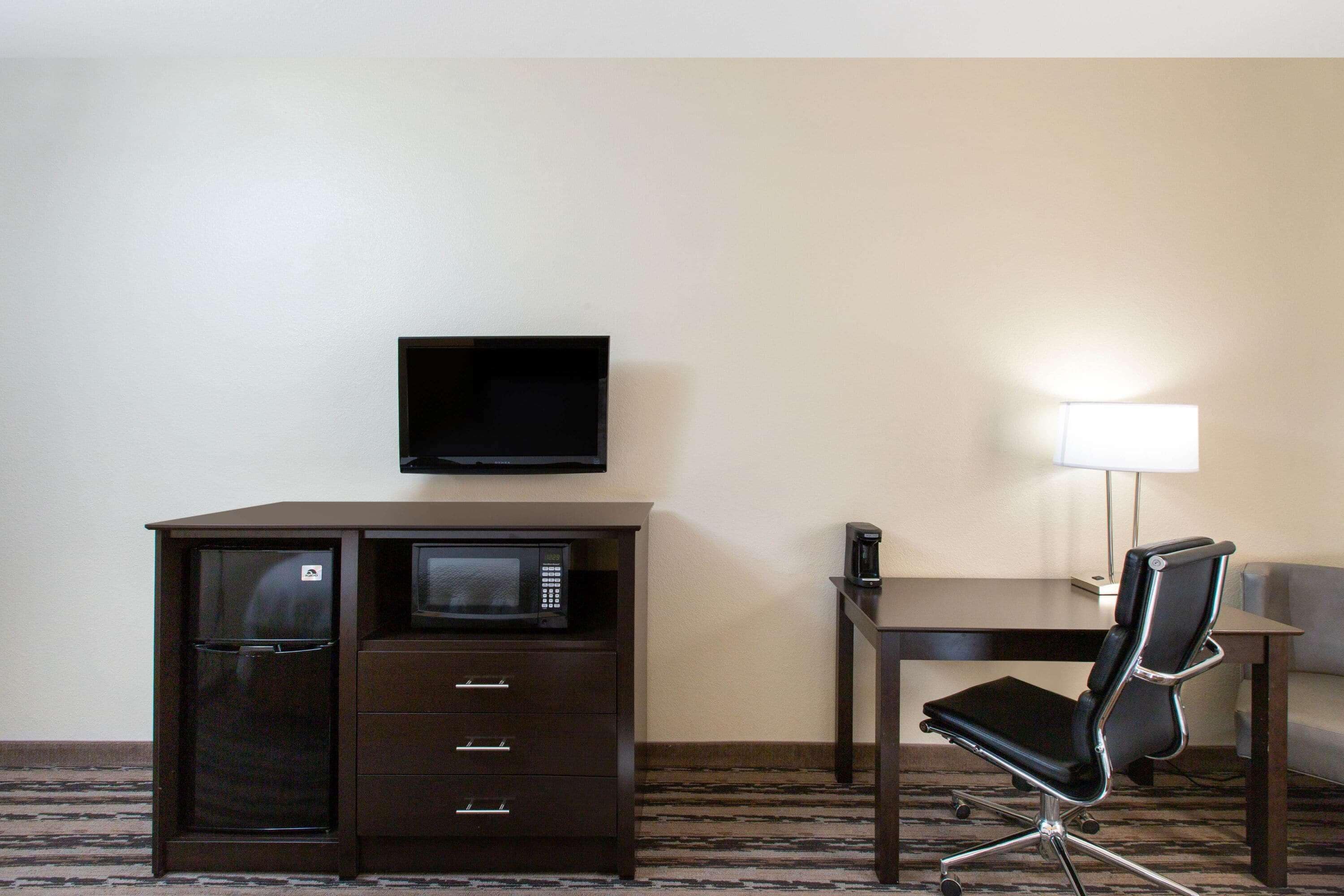 Ein Hotelzimmer mit dunklem Holztisch, schwarzem Bürostuhl, flachbildschirmfernseher über dem Tisch, einem kleinen Tisch mit Lampe und einer Minibar.