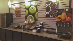 A hotel reception area with a coffee station, a fruit juice dispenser, and a display of pastries and donuts. - La Quinta Inn by Wyndham Lincoln, Nebraska (Lincoln)