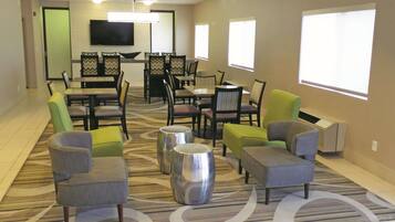 A hotel lobby with a dining area, seating arrangements, and a flat-screen TV mounted on the wall.