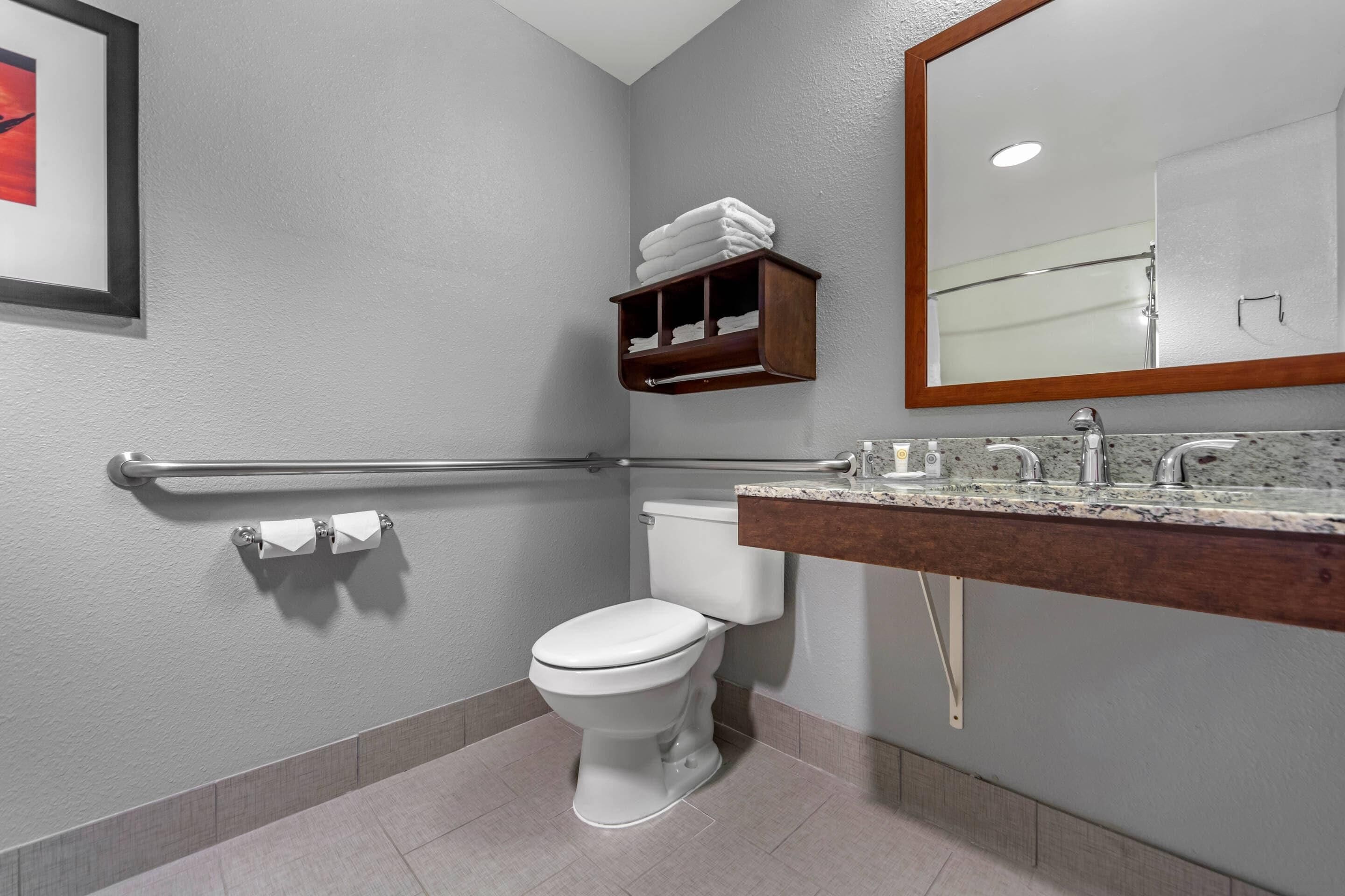 Un baño moderno con inodoro, lavabo y espejo. Hay un perchero sobre el inodoro y un pasamanos en la pared.
