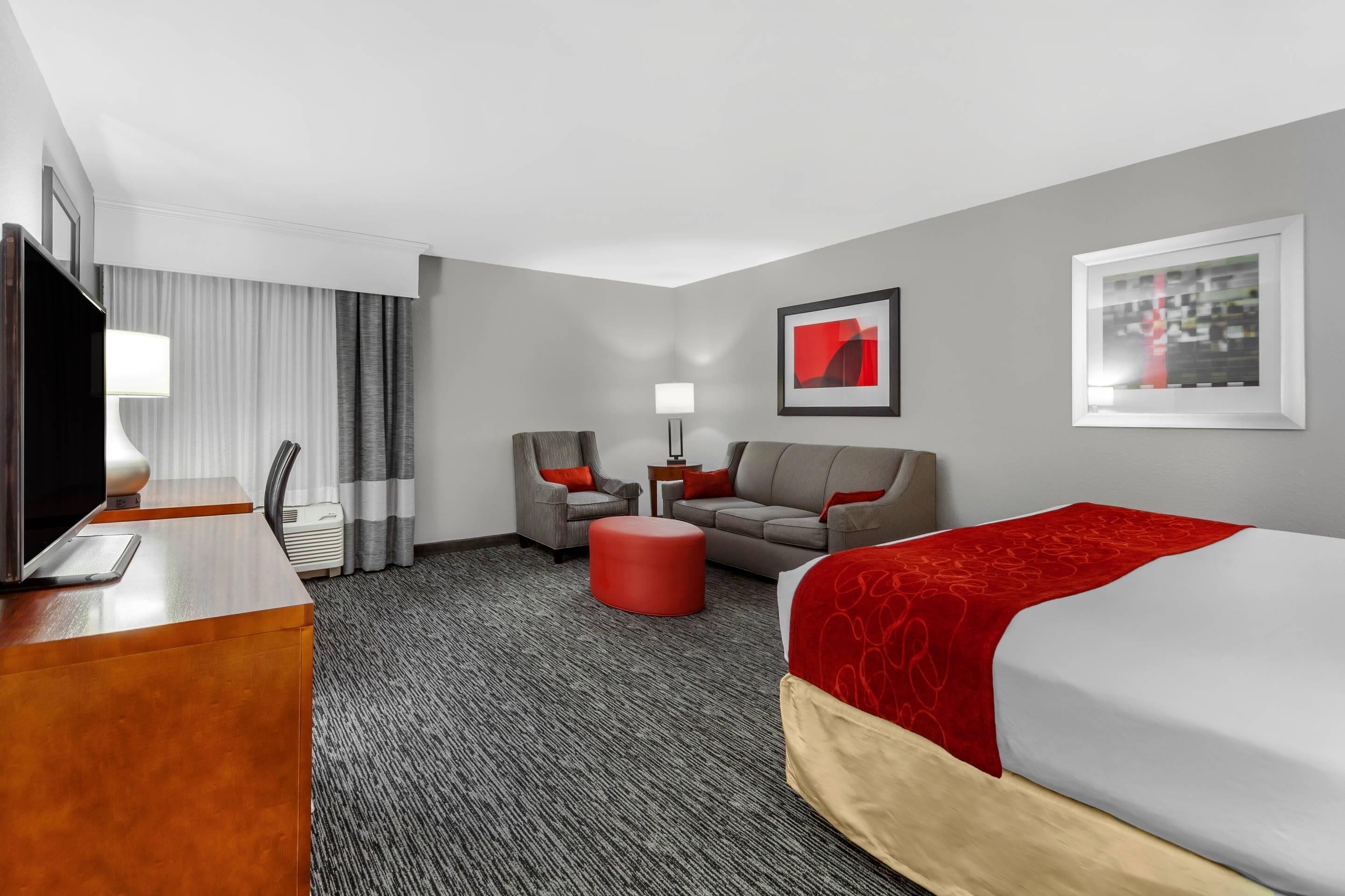 A hotel room with a bed, a desk with a TV, a sofa, a red ottoman, and a chair.