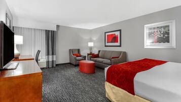 A hotel room with a bed, a desk with a TV, a sofa, a red ottoman, and a chair.