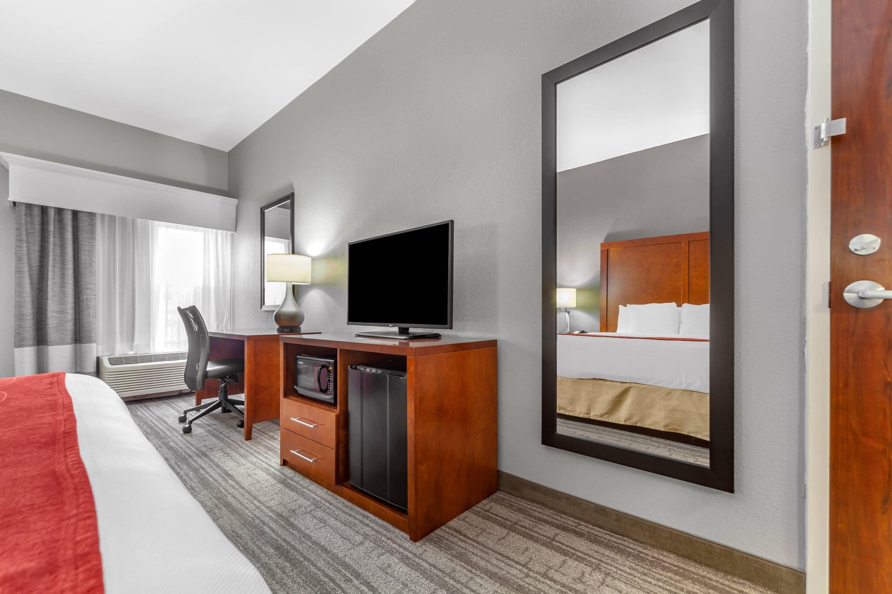 A hotel room with a large bed, a desk with a television, a mirror, a wardrobe, and a view of the hotel room through the mirror.