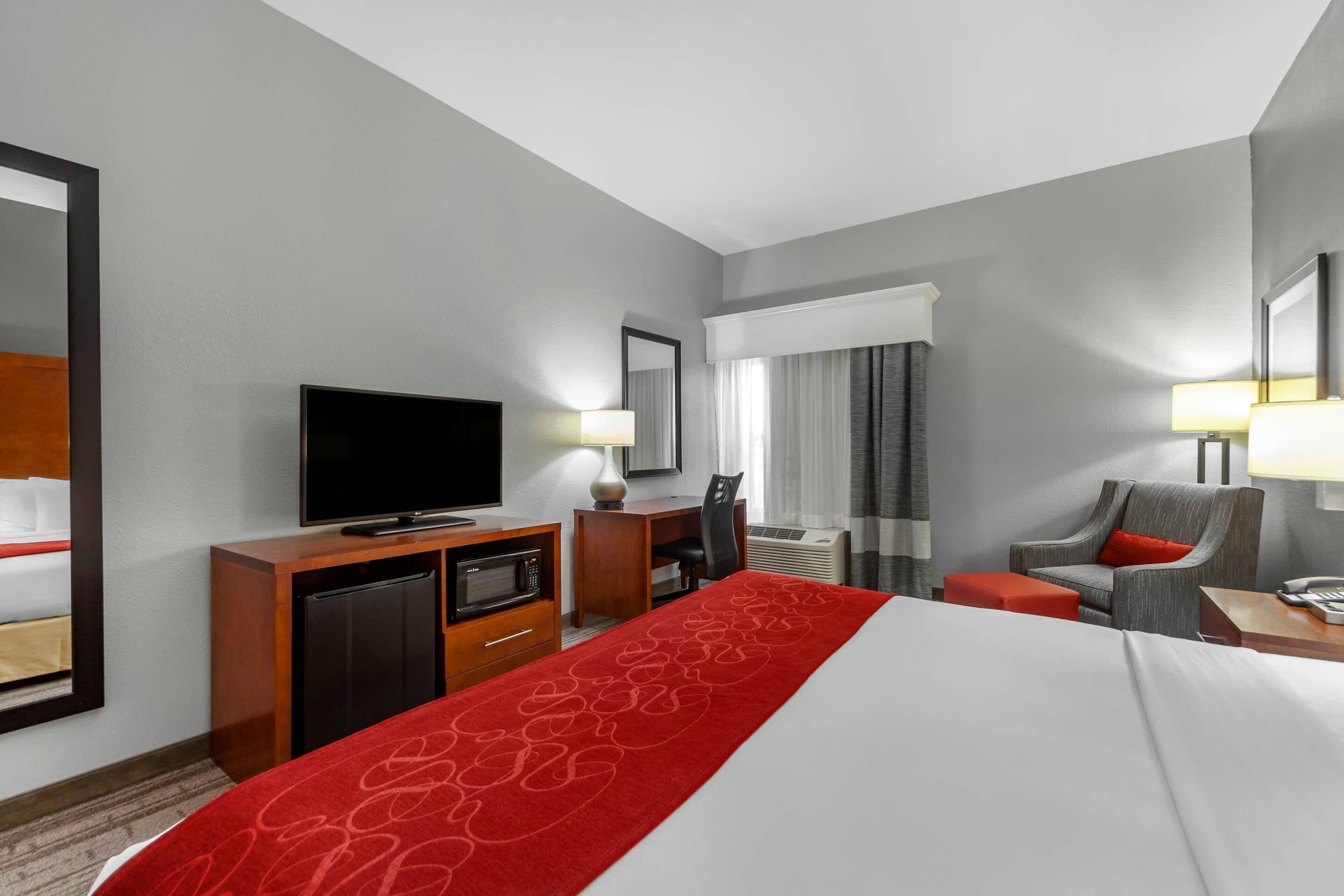 Habitación de hotel con cama, escritorio, televisor, silla y una mesita de noche con lámpara.
