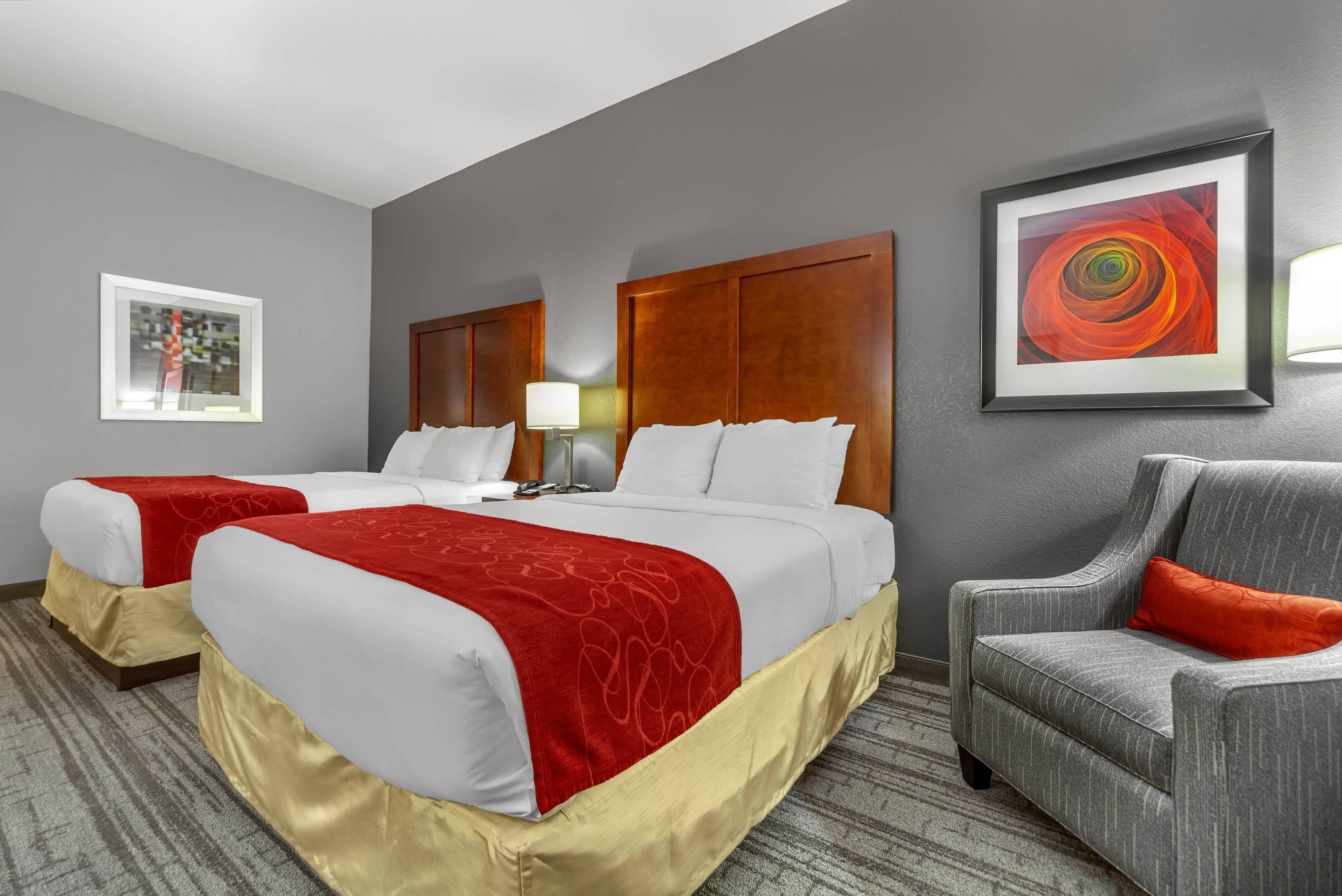 A hotel room with two beds, a gray armchair, a framed abstract painting, and a lamp.
