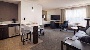 Una habitación de hotel moderna con kitchenette, zona de comedor, sofá, televisor y un ventanal con cortinas.