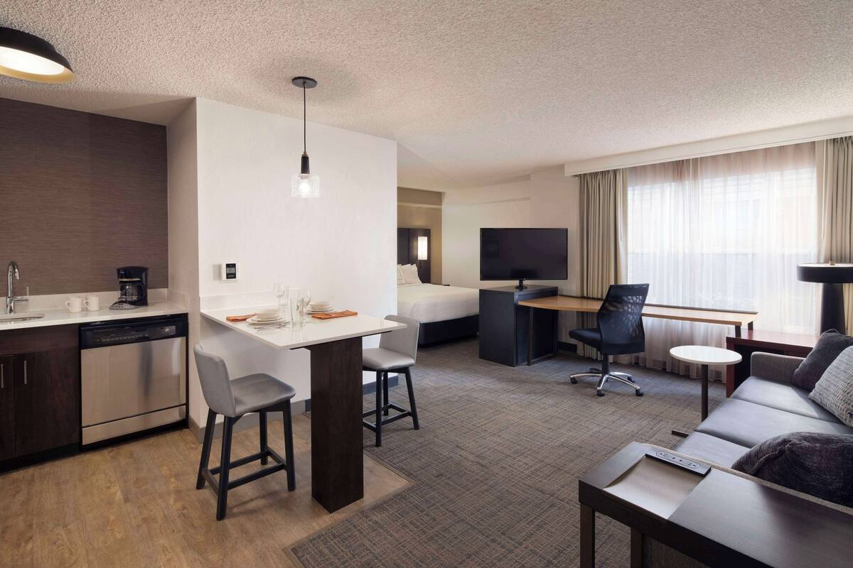 A modern hotel room with a kitchenette, a dining area, a sofa, a TV, and a large window with curtains.