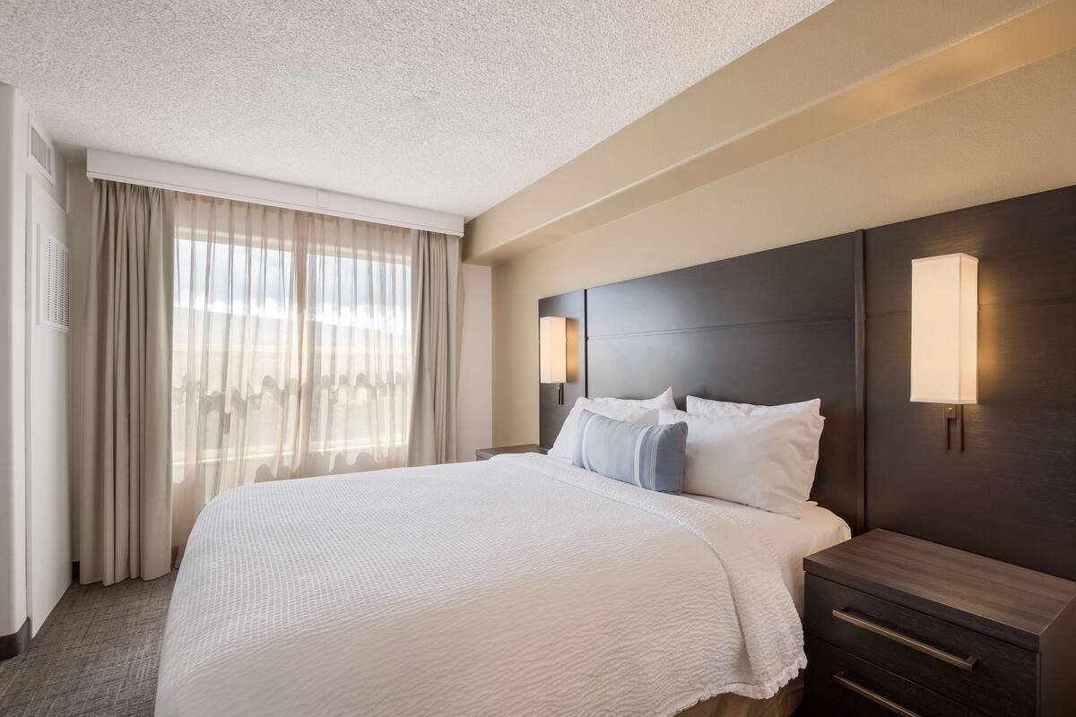 A hotel room with a large bed, a dark headboard, two bedside lamps, and a nightstand with drawers.