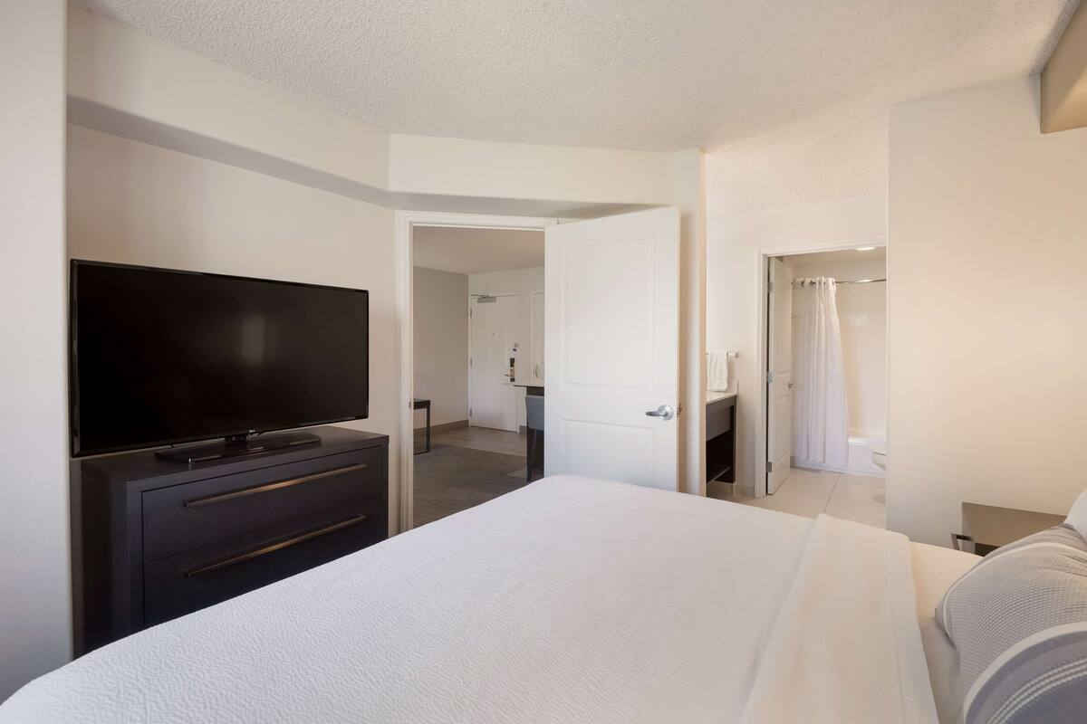 A hotel room with a large bed, a flat-screen TV on a dresser, a bathroom with a shower curtain, and a door leading to another room.