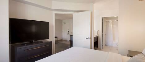 Una habitación de hotel con una cama grande, un televisor de pantalla plana sobre un tocador, un baño con cortina de ducha y una puerta que da a otra habitación.