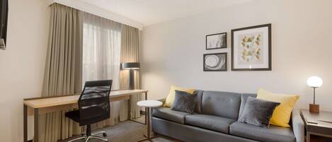 A hotel room with a desk, a grey sofa, two pillows, a lamp, and framed artwork on the wall.