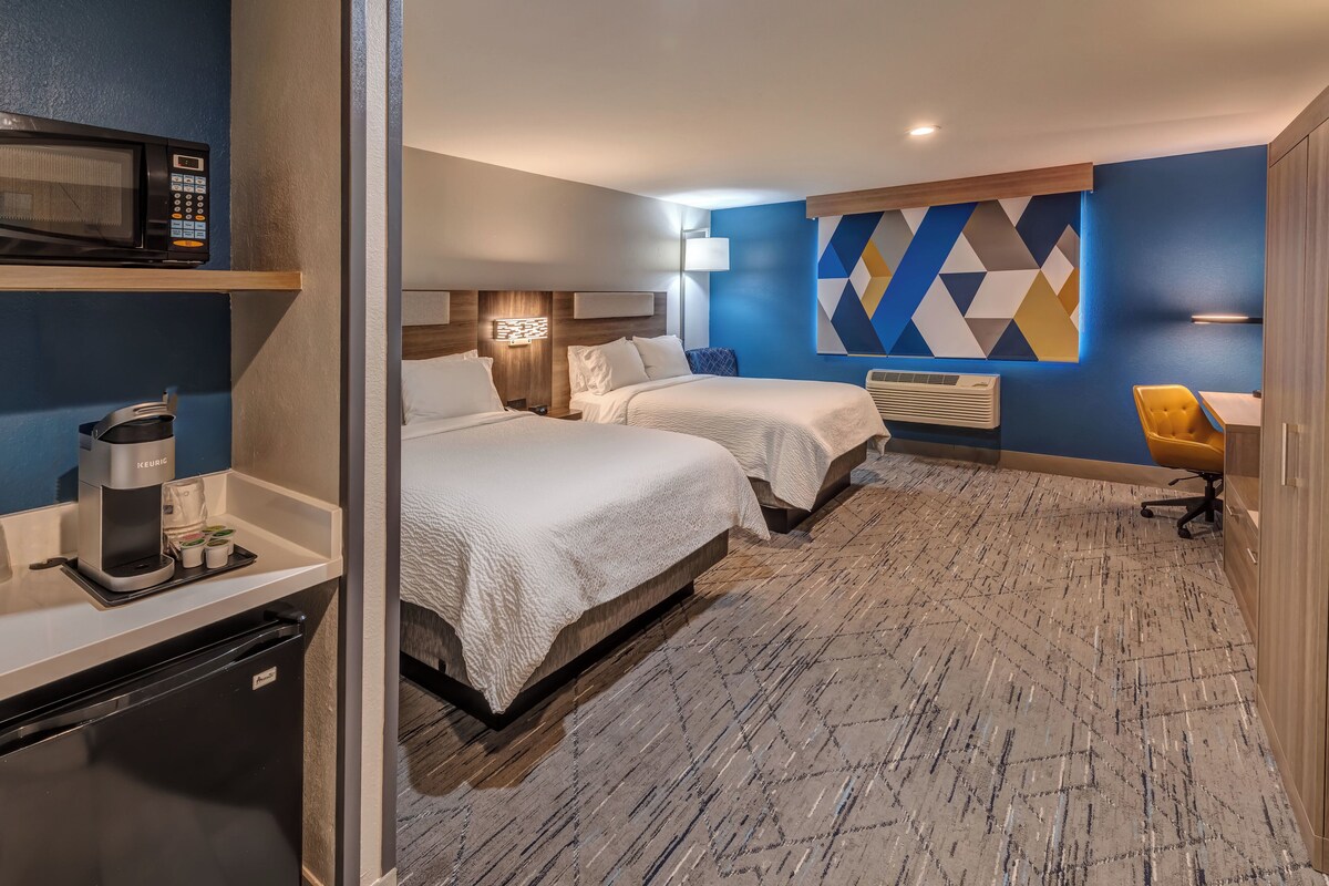 Une chambre d’hôtel avec deux lits, un minibar, une machine à café, un bureau avec une chaise et une décoration murale aux motifs géométriques.