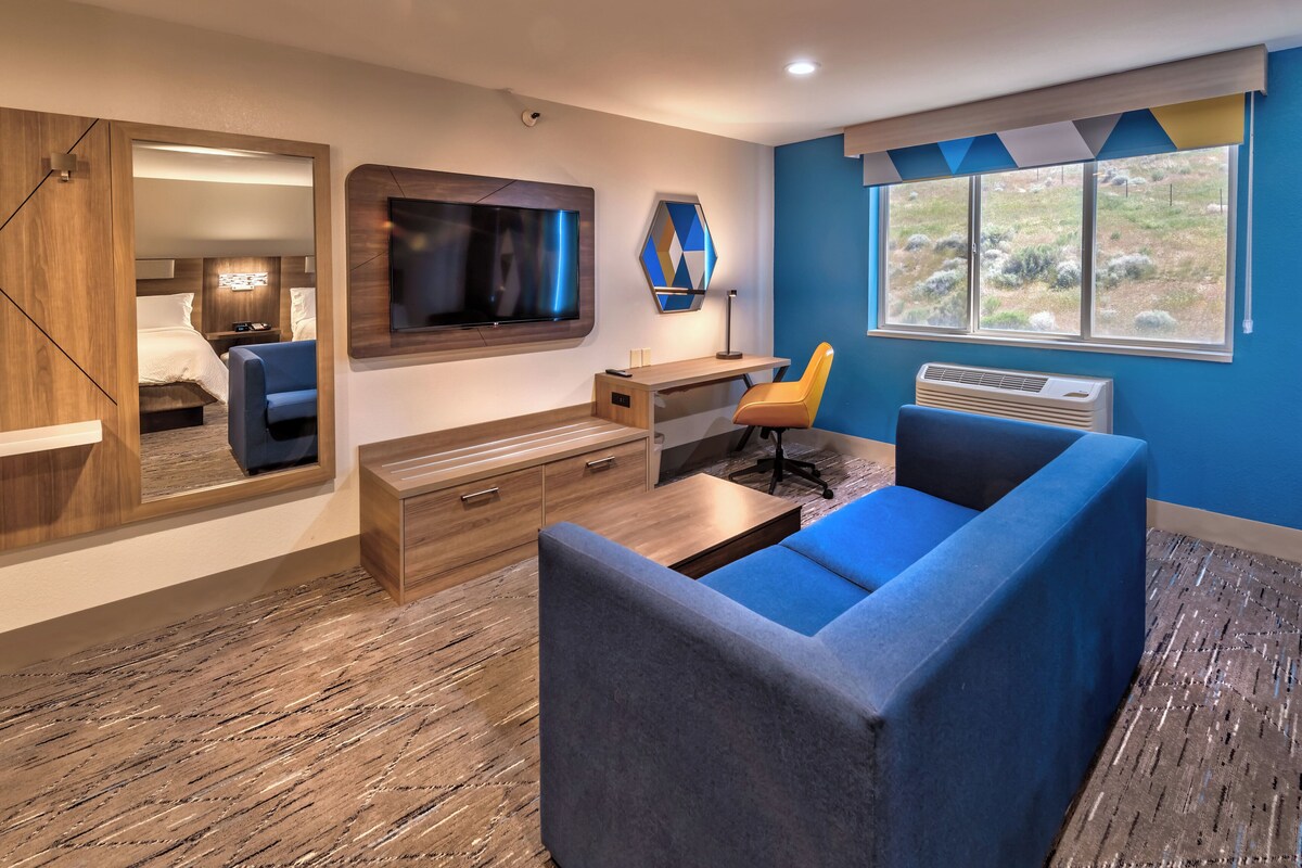 Une chambre moderne dotée d’un grand téléviseur à écran plat fixé au mur, d’un bureau en bois avec des tiroirs, d’un canapé bleu et d’une fenêtre donnant sur l’extérieur.