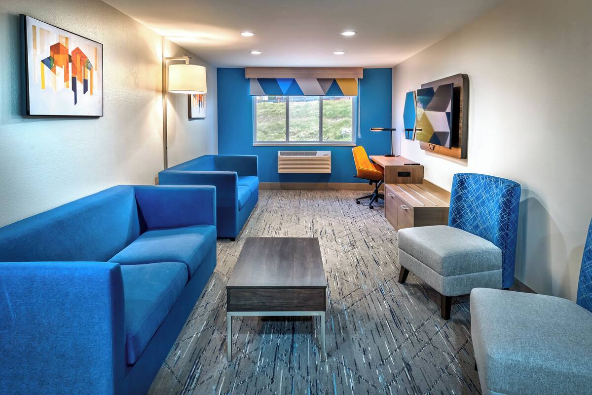A modern hotel room with blue sofas, a wooden coffee table, and a desk with an orange chair.