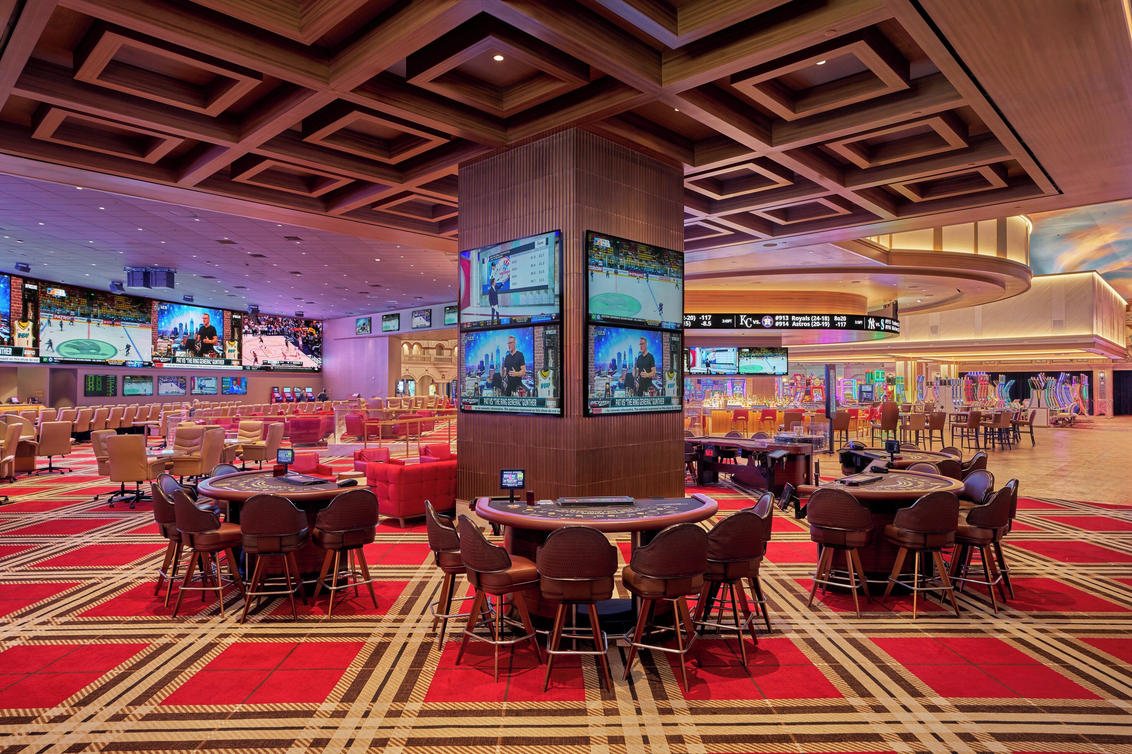 Een casino met meerdere speeltafels en stoelen, een centraal pilaar met schermen, en een rode tapijtvloer.