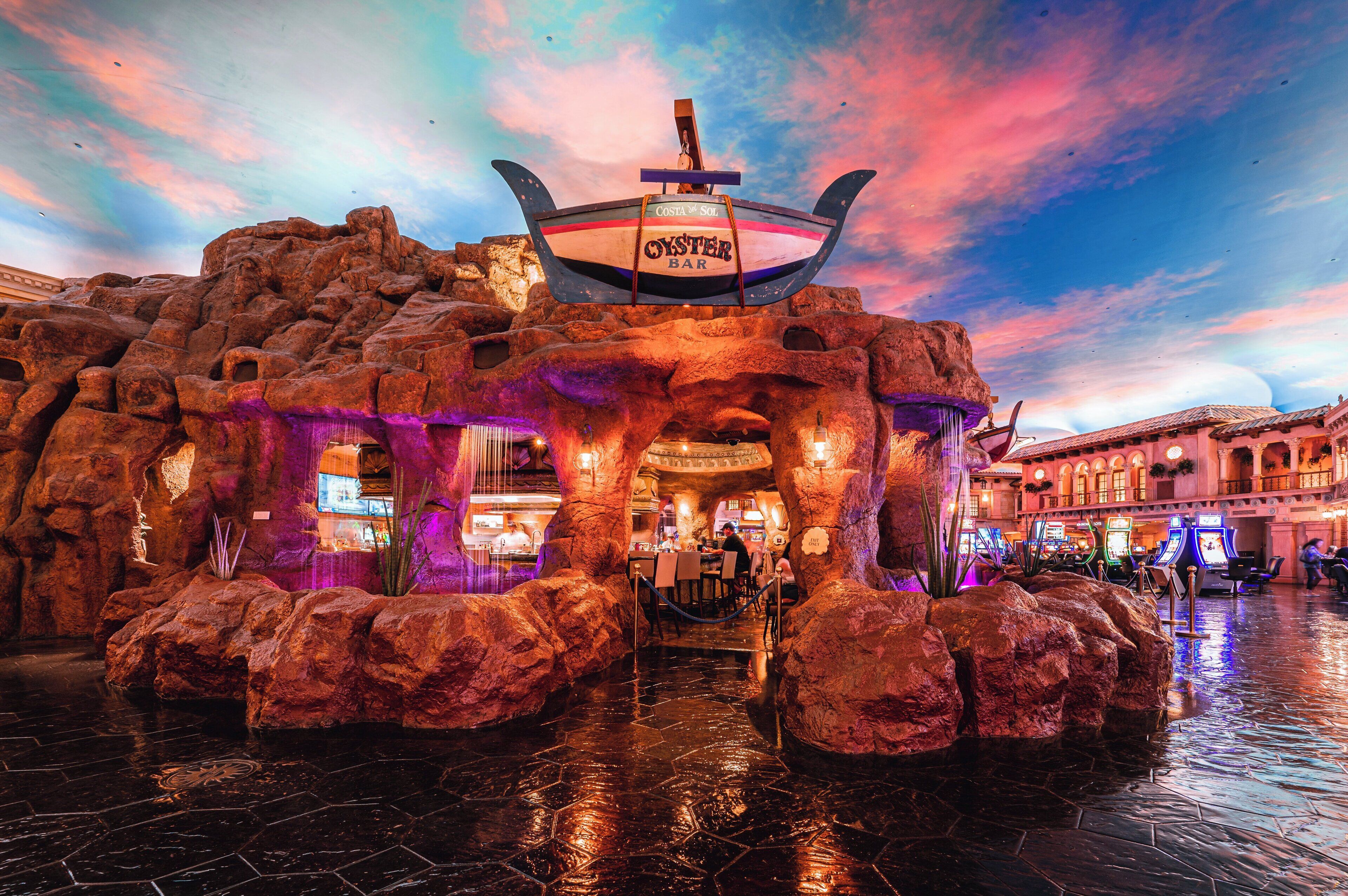 Un bar llamado "Oyster Bar" con un letrero con forma de barco, ubicado en una zona rocosa interior con vista a un edificio al fondo.