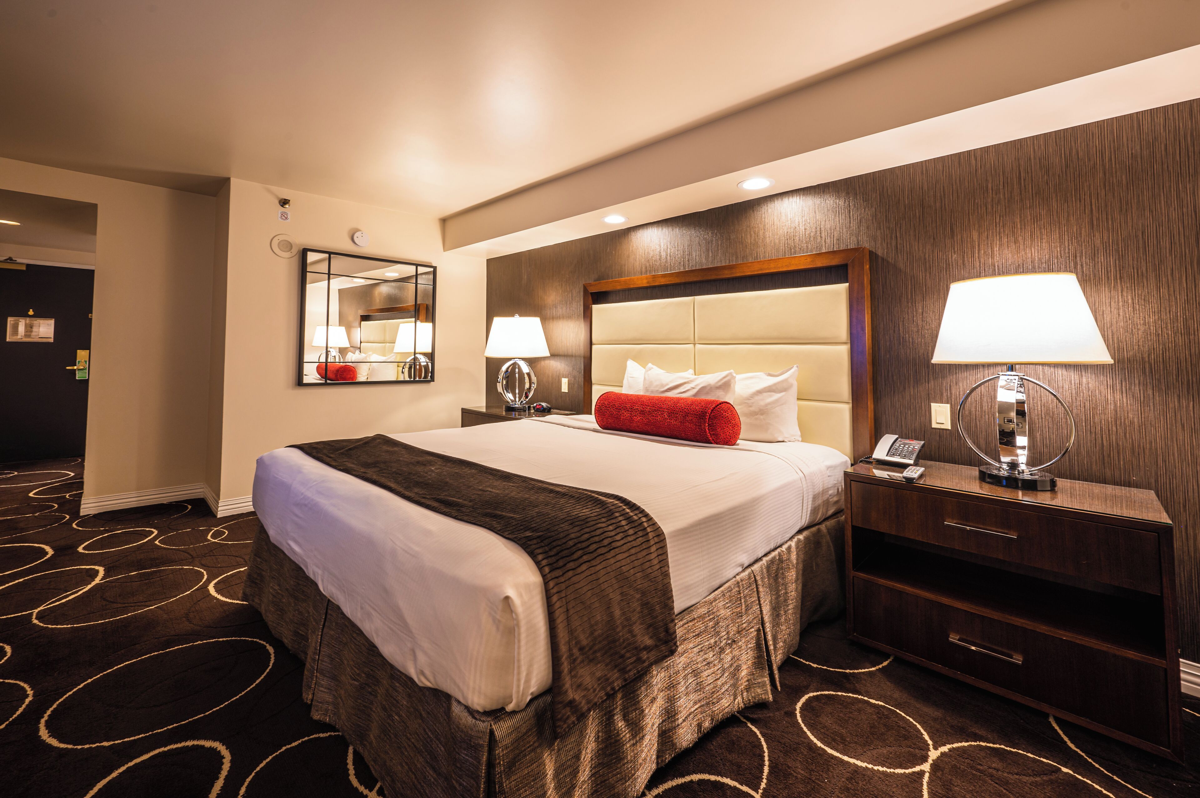 A hotel room with a large bed, two bedside tables with lamps, a wooden headboard, a mirror, and a carpet with a circular pattern.