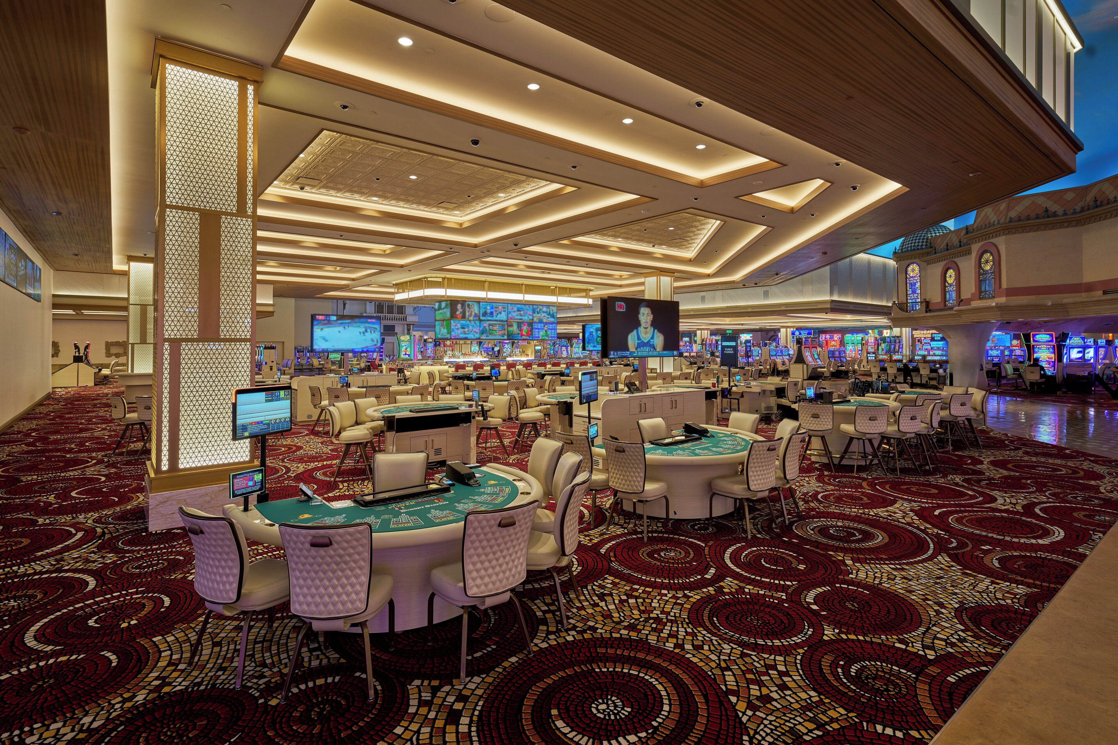 Un casino espacioso con múltiples mesas y sillas de juego, una pantalla grande mostrando un juego y una alfombra estampada.