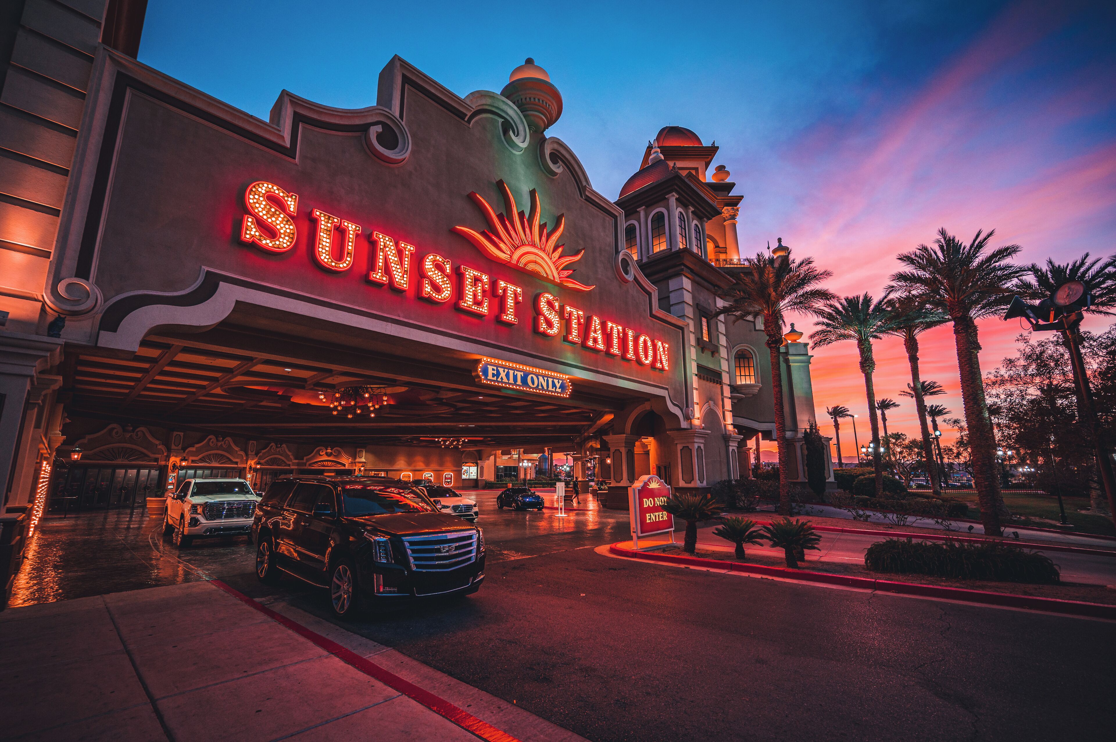 L’extérieur de l’hôtel Sunset Station, avec son grand panneau lumineux, son entrée abritée et son parking où sont garées plusieurs voitures.