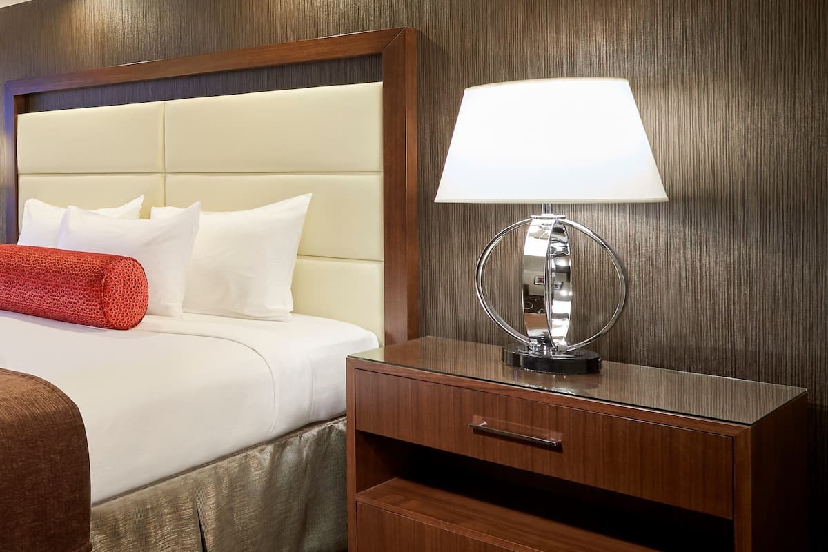 Ein ordentlich gemachtes Bett mit weißen Leinentüchern und einem roten Zierkissen, ein Nachttisch mit Lampe und ein hölzernes Kopfende.