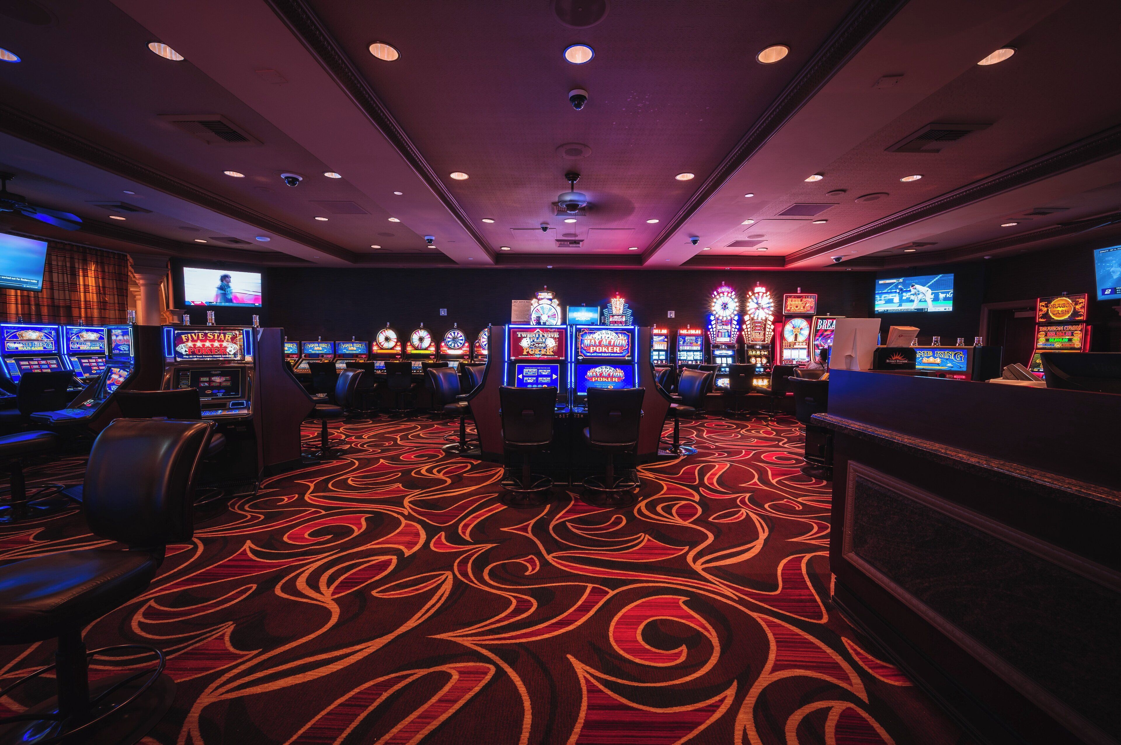 Une salle de casino avec de nombreux appareils à sous, une moquette à motifs et des caméras de surveillance au plafond.