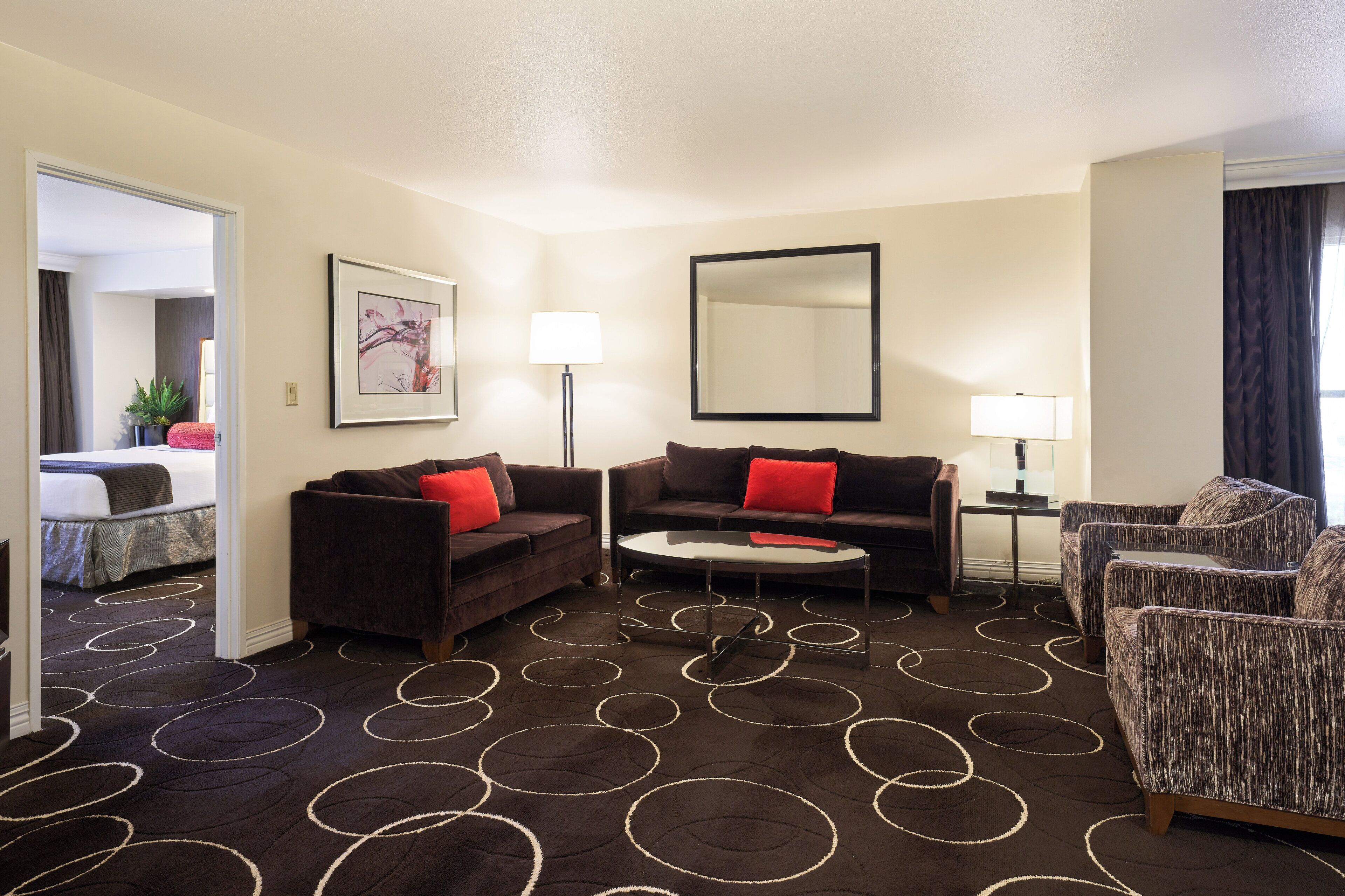 A hotel room with a dark patterned carpet, a sofa, a chair, a small table, and a bed in the background.