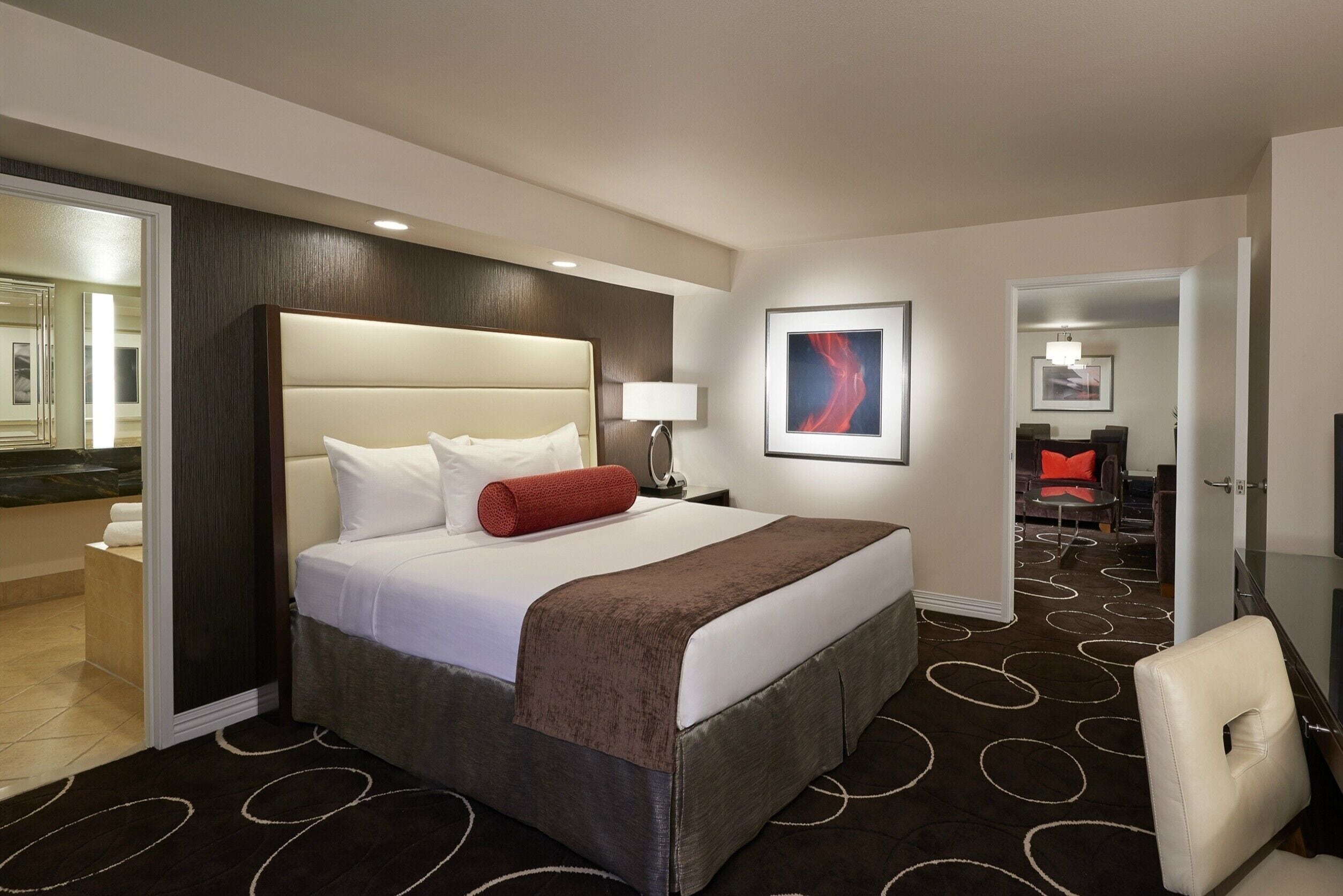 A modern hotel room with a large bed, a headboard, a bedside table with a lamp, a red pillow, a brown throw blanket, and a chair.