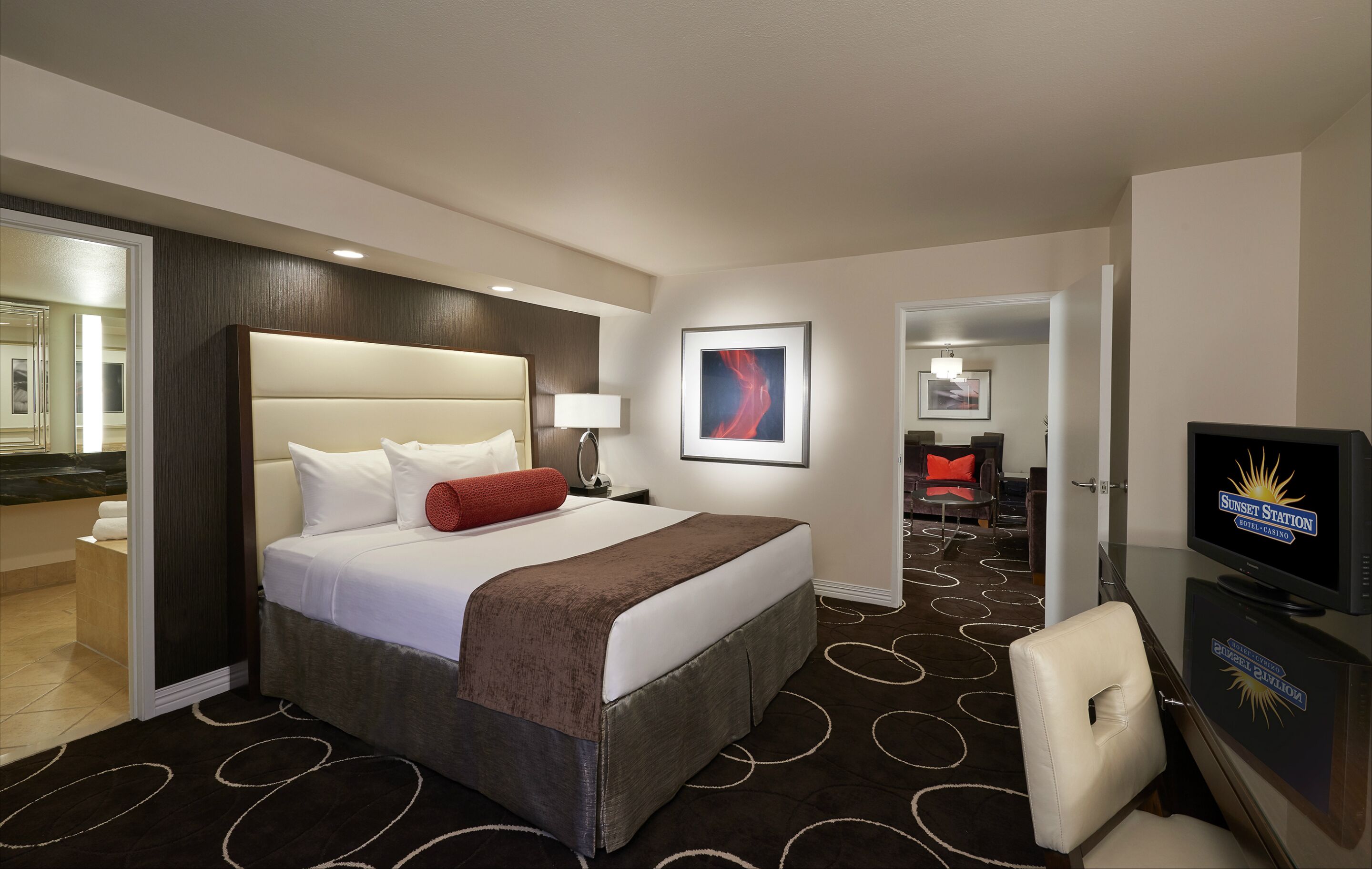 A hotel room with a large bed, a television, a desk with a chair, and a bathroom visible through an open door.