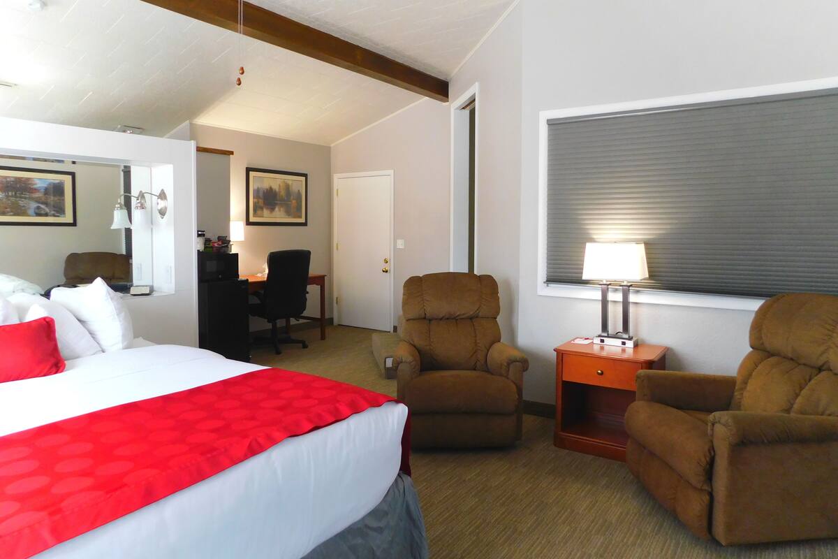 Une chambre d’hôtel comprenant un lit, deux fauteuils, un bureau et une fenêtre avec des stores.