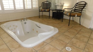 Un bagno con una grande vasca, due sedie, un tavolino con un cesto e una finestra con persiane.