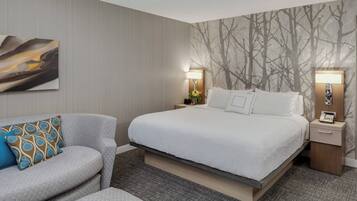Habitación de hotel con cama, sofá, dos mesitas de noche con lámparas, y pared con estampado de árbol.