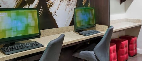 A workspace with two desks, each with a computer monitor and keyboard. There are two office chairs and a red ottoman. A large painting is on the wall behind the desks.