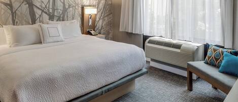 A hotel room with a large bed, a bedside table with a lamp, a bench with cushions, and a window with curtains.
