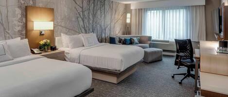 Habitación de hotel con dos camas, un escritorio, una silla, una lámpara y un ventanal grande con cortinas.