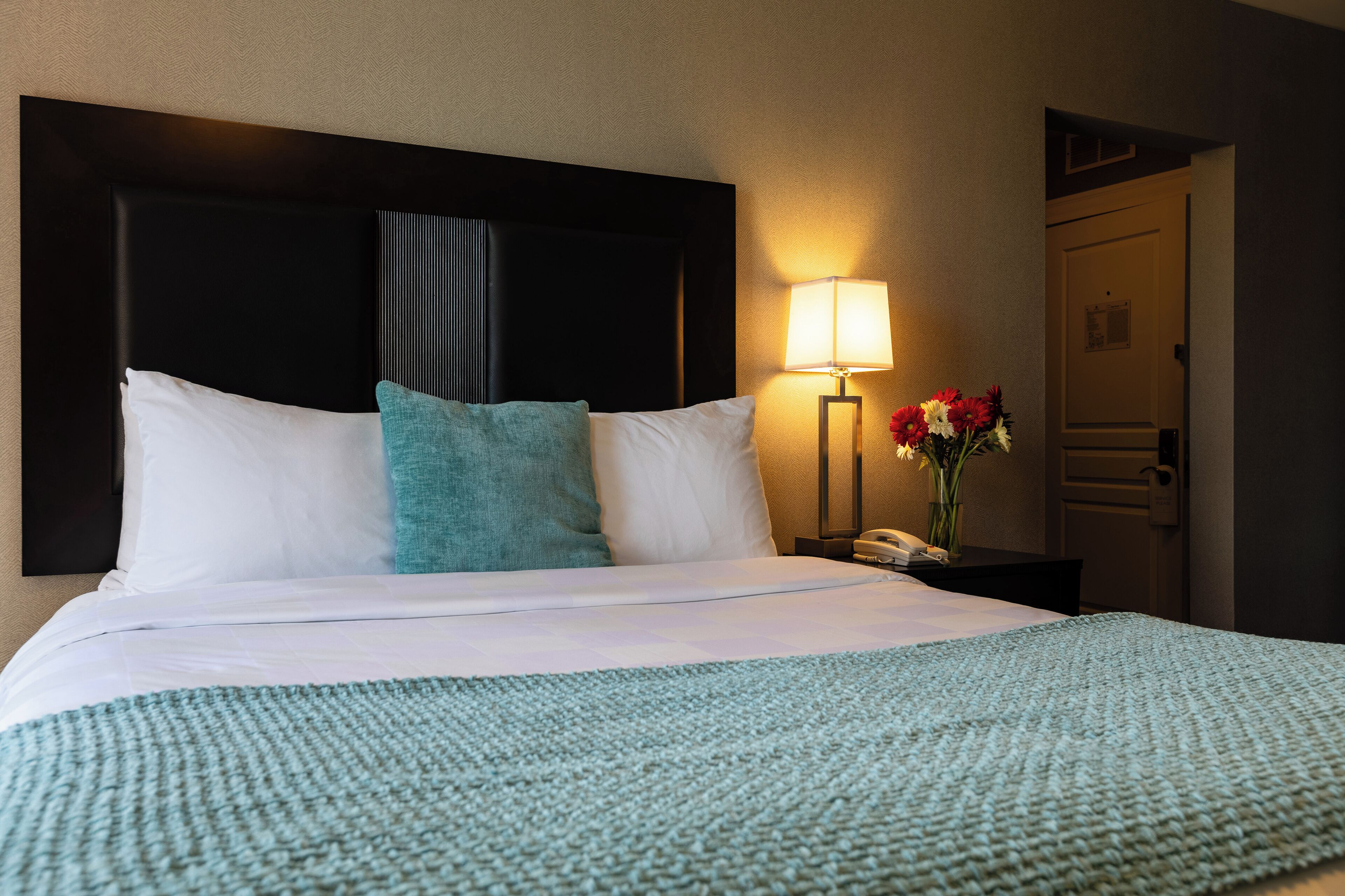 Una habitación de hotel con una cama grande, una mesita de noche con lámpara y una puerta.
