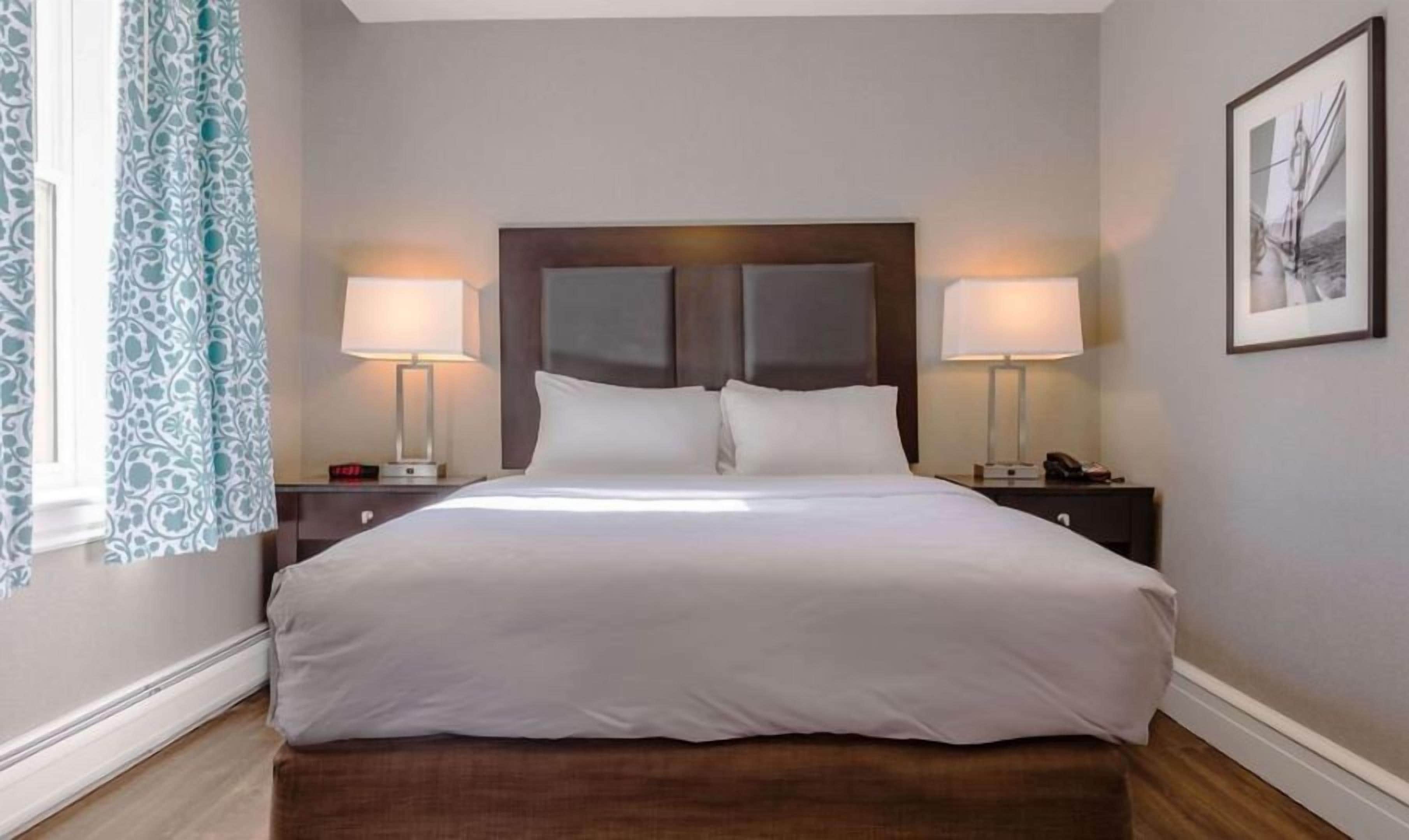 Una habitación de hotel con una cama grande, dos mesitas de noche, una lámpara y una ventana con cortinas, además de un cuadro en la pared.