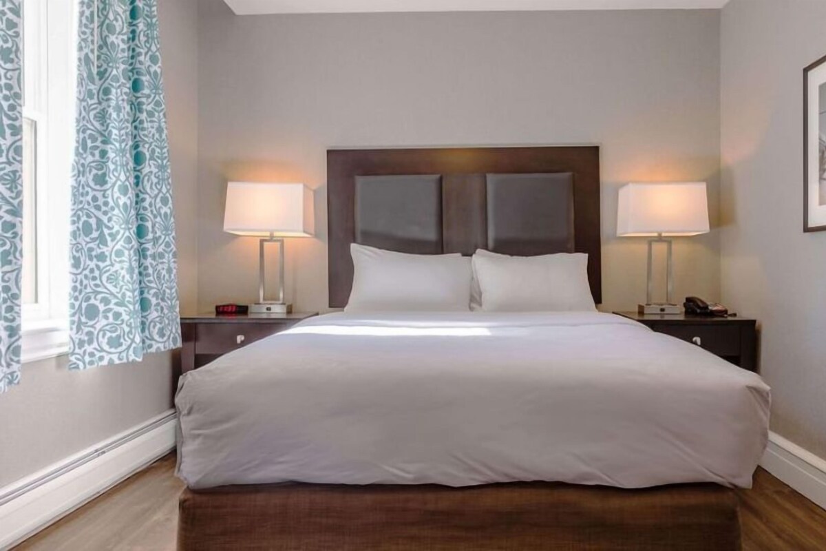 Una habitación de hotel con una cama grande, dos mesitas de noche, una lámpara y una ventana con cortinas, además de un cuadro en la pared.