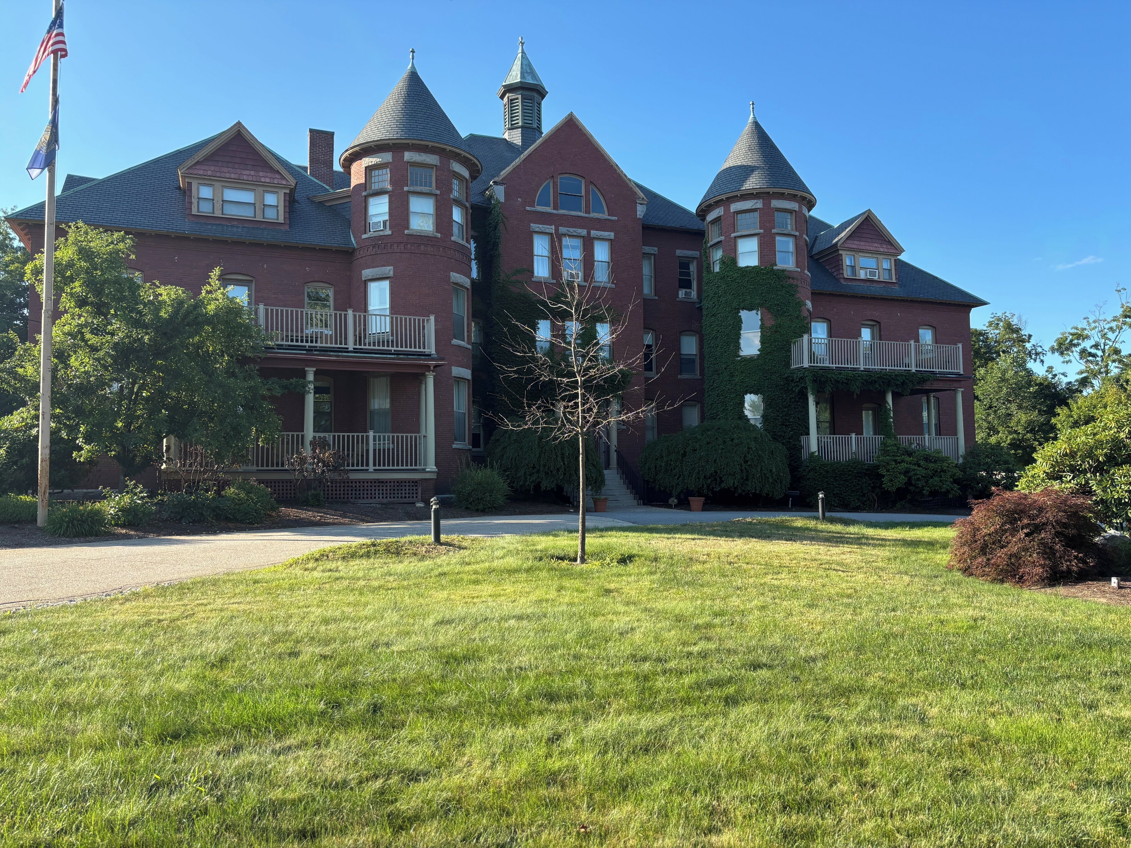A large, multi-story brick building with a central tower and two smaller towers, surrounded by well-maintained lawns and a flagpole.